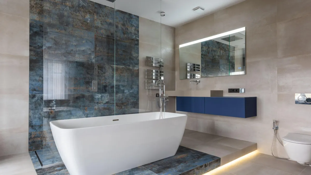 Contemporary bathroom with a white freestanding bathtub and floor-mounted faucet, set against a striking feature wall of blue distressed tiles. Beige tiles cover the rest of the room, complemented by a sleek blue vanity unit with a large mirror above. Ambient lighting enhances the serene and modern atmosphere of the space