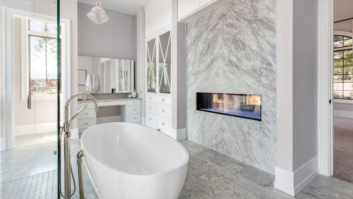 Luxurious bathroom with marble flooring and walls, featuring a standalone white tub and a wall-mounted fireplace. The room boasts a double vanity with shaker-style cabinets and a large mirror. Crystal lighting adds a touch of sophistication, while a large window bathes the space in natural light, showcasing the elegant and tranquil setting.