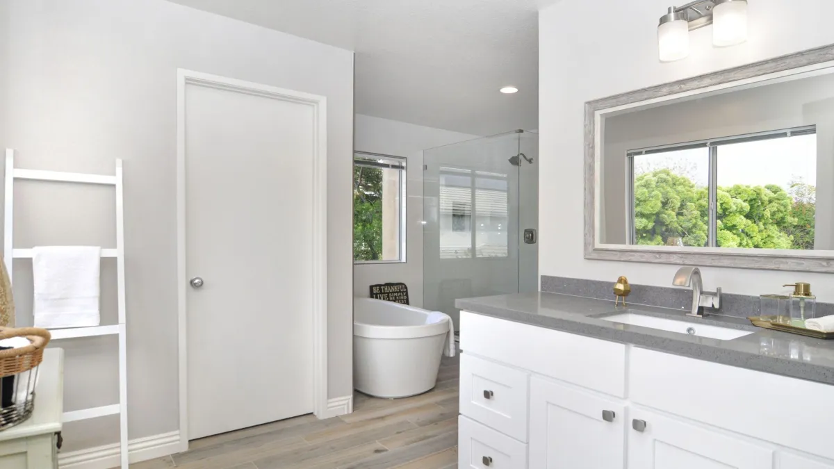 Bright and airy bathroom with a minimalist design, featuring a large mirror over a white vanity with a grey countertop and modern fixtures. A freestanding tub is visible through the doorway, complemented by soft natural light, a white towel ladder, and greenery visible through the window, creating a tranquil spa-like atmosphere.