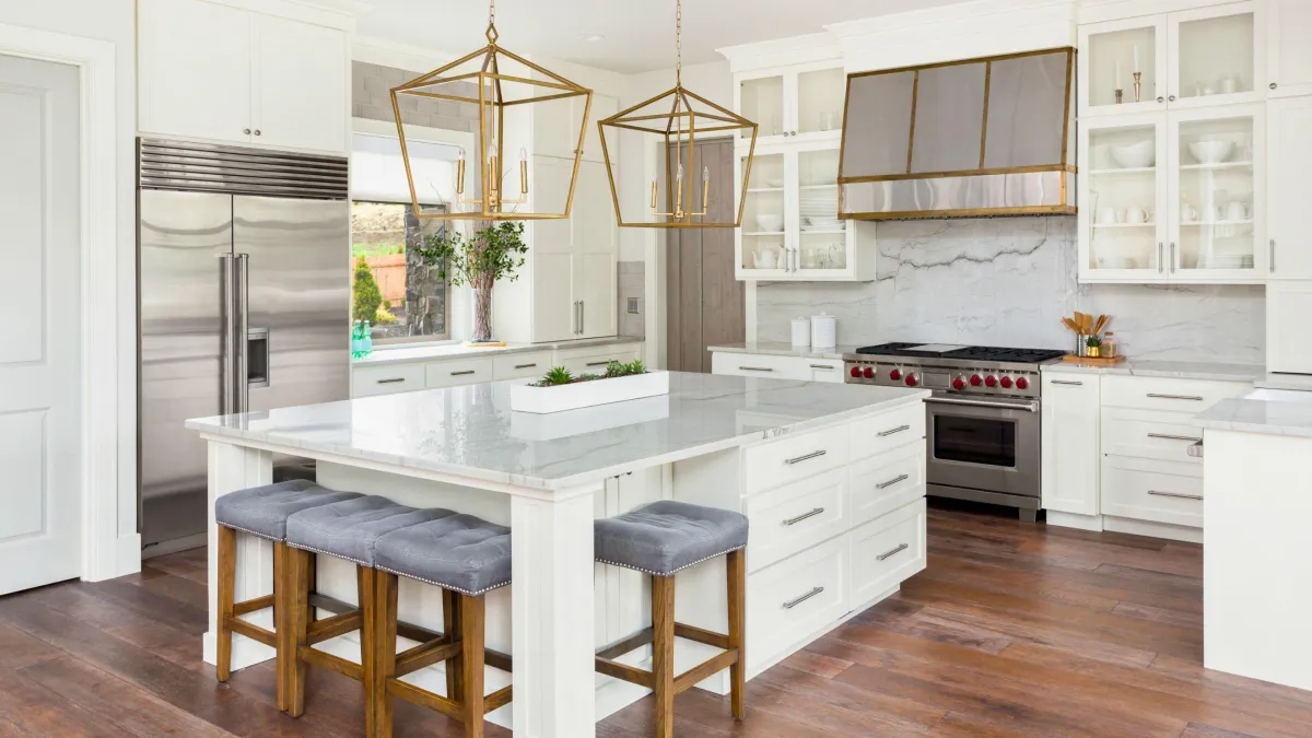 This traditional kitchen exudes elegance with its custom white cabinetry and luxurious marble countertops. The large island at the center, equipped with a built-in sink and stylish gray upholstered stools, invites casual dining and conversation. Statement geometric pendant lights in a brushed gold finish hover above, casting a warm glow and adding a touch of sophistication. High-end stainless steel appliances, including a range with red knobs, offer a pop of color and hint at professional-grade culinary capabilities. The rich, dark wood flooring contrasts beautifully with the bright cabinetry, creating a welcoming and balanced aesthetic. Glass-fronted cabinets provide an opportunity to display fine dishware, completing this kitchen's blend of classic style and modern functionality.