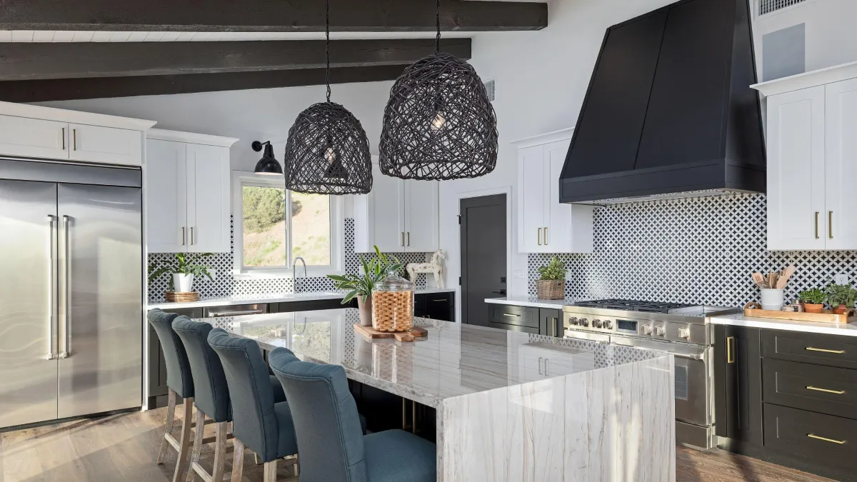 This kitchen strikes a stunning balance of modern design and natural texture. The waterfall marble island is a statement piece, complemented by plush teal bar stools, which add a pop of color to the neutral palette. Above, unique woven pendant lights bring an organic touch, contrasting with the sleek lines of the white cabinetry and dark, dramatic range hood. The geometric patterned backsplash infuses the space with personality, while the stainless steel appliances offer a glint of modernity and efficiency. The open beams overhead add architectural interest, harmonizing with the herringbone wood flooring. It's a space that's equally prepared for casual family meals or entertaining guests in style.