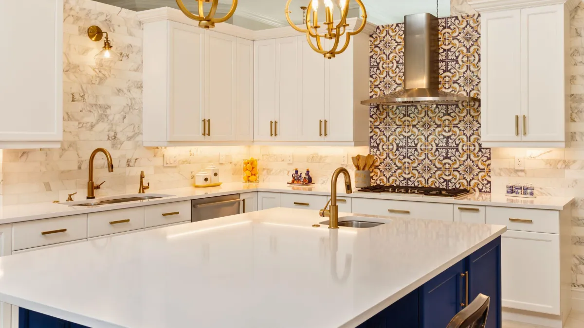 This kitchen design perfectly marries classic elegance with bohemian flair, presenting a bright and airy space adorned with a luxurious marble backsplash. The deep blue lower cabinets add a rich pop of color, beautifully contrasted by the crisp white upper cabinets and countertops. Gold accents from the cabinet handles to the faucet bring a touch of warmth and sophistication, harmonizing with the ornate, colorful tiles that create a stunning focal point above the range. This kitchen not only stands out for its bold use of pattern and color but also exemplifies a space where cooking and style come together harmoniously.
