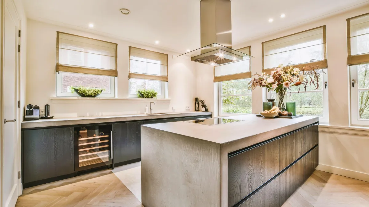  This kitchen blends urban modernity with natural elements, presenting a spacious area highlighted by a striking concrete island at its heart. The sleek black cabinetry with wood accents creates a sophisticated look, while the built-in wine cooler adds a touch of luxury. Large windows dressed with chic roman shades allow natural light to illuminate the space, enhancing the fresh greenery on the windowsills. The herring herringbone wood floor complements the industrial feel of the concrete, and the statement extractor hood above the island ensures a fusion of form and function. This kitchen is a perfect canvas for culinary exploration and social gatherings, reflecting a contemporary lifestyle with its design-forward approach.