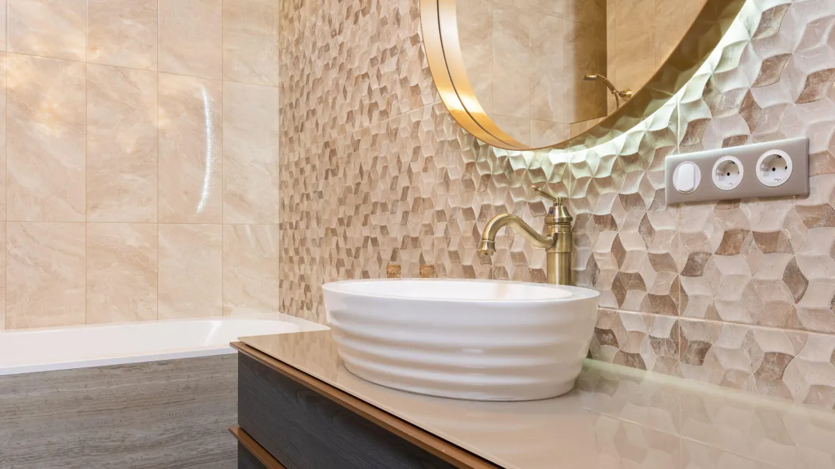 Elegant bathroom detail showcasing an artisanal white vessel sink on a marble countertop with a brushed gold faucet, set against a textured wall with a unique 3D wave pattern in neutral tones. A round mirror with a golden frame adds a touch of luxury, reflecting the intricate design of the wall and the warm lighting that enhances the sink area's sophisticated and artistic ambiance.