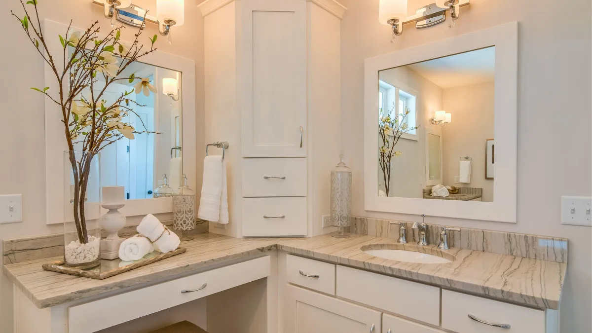 Inviting bathroom with a warm and cozy feel, showcasing custom white cabinetry and a built-in vanity with a tan marble countertop. A large framed mirror reflects a vase with delicate branches, adding a touch of nature. Elegant glass canisters and neatly rolled towels create a spa-like atmosphere, with soft lighting from wall sconces enhancing the serene environment.