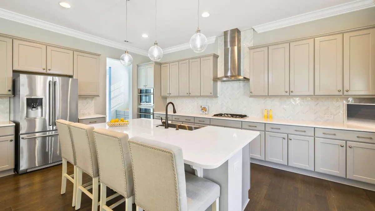  This image depicts a bright and luxurious kitchen, boasting custom cabinetry in a soft taupe that exudes warmth and sophistication. The large central island, topped with a pristine white countertop, serves as a focal point and a casual dining spot with its elegant high-back chairs. Unique herringbone-patterned backsplash tiles add depth and texture to the design, while the modern stainless steel appliances offer functionality and style. Above, delicate pendant lights hang gracefully, contributing to the kitchen's overall serene and inviting atmosphere. Rich, dark wood flooring anchors the space, providing a lovely contrast to the light cabinetry. This kitchen elegantly combines traditional elements with modern finishes to create a space that is both welcoming and refined.