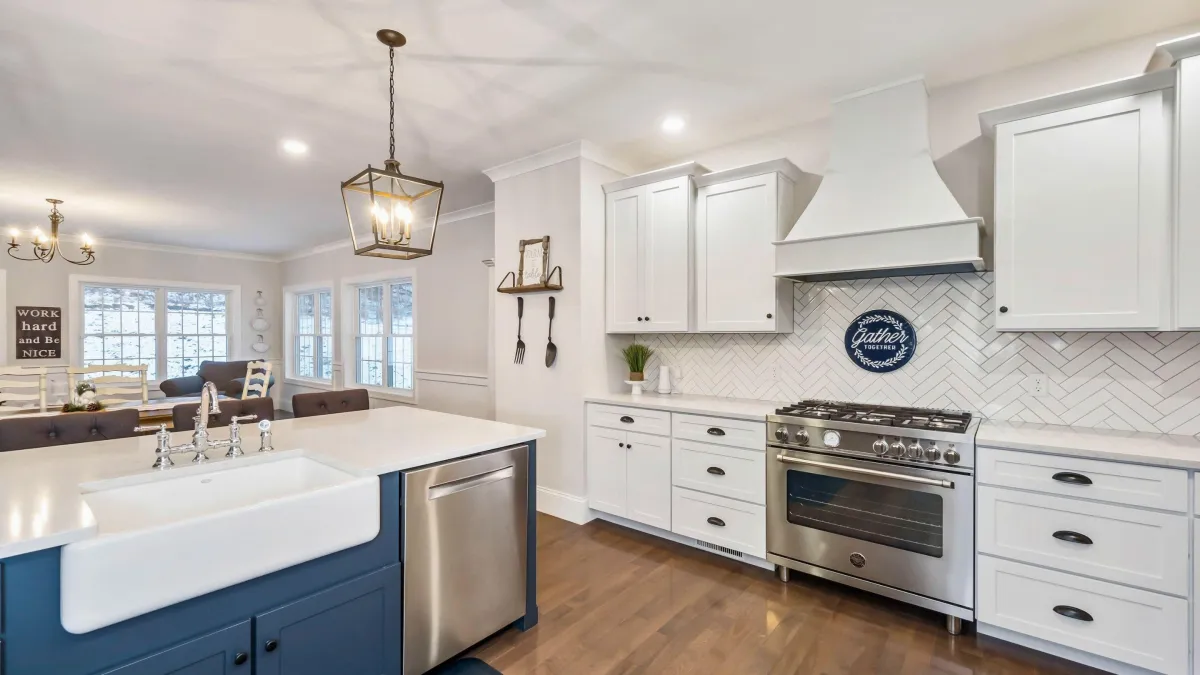  This inviting kitchen design showcases the beauty of traditional craftsmanship combined with modern elegance. The classic white shaker cabinetry is paired with a rich blue island base, making a statement with their clean lines and timeless appeal. A stylish farmhouse sink takes center stage, complemented by high-end stainless steel appliances, including a professional-grade range. The herringbone tile backsplash adds texture and interest, while the sophisticated pendant lighting fixtures cast a warm and welcoming glow. The space is tied together with hardwood flooring, creating a seamless flow into the cozy living area. Decorative touches like the "Gather Together" sign add a personal and homely feel, emphasizing this kitchen's role as the heart of the home.