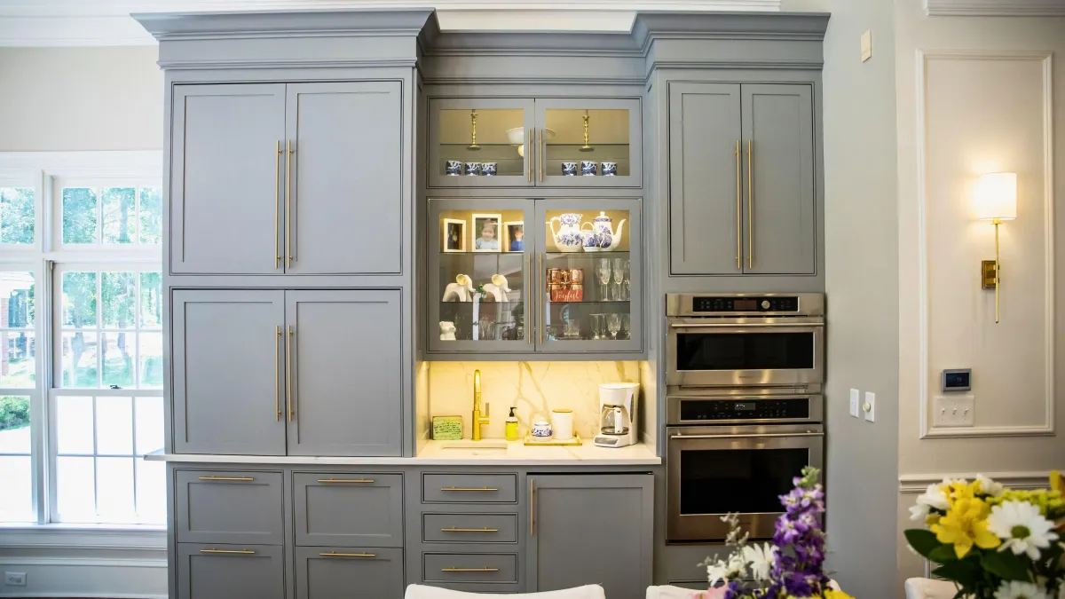 This kitchen strikes a perfect balance between classic charm and modern sophistication, featuring soft gray custom cabinets with elegant gold hardware. The glass-front upper cabinets offer a display for fine chinaware, adding a personal touch to the space. Integrated high-end appliances provide a seamless look and state-of-the-art functionality, ideal for the culinary enthusiast. Under-cabinet lighting casts a warm glow on the marble backsplash, enhancing the ambiance of the room. This kitchen's open concept is bathed in natural light from the large windows, creating a welcoming space for both cooking and gathering. With its thoughtful design and attention to detail, this kitchen is a testament to timeless style meeting modern convenience.