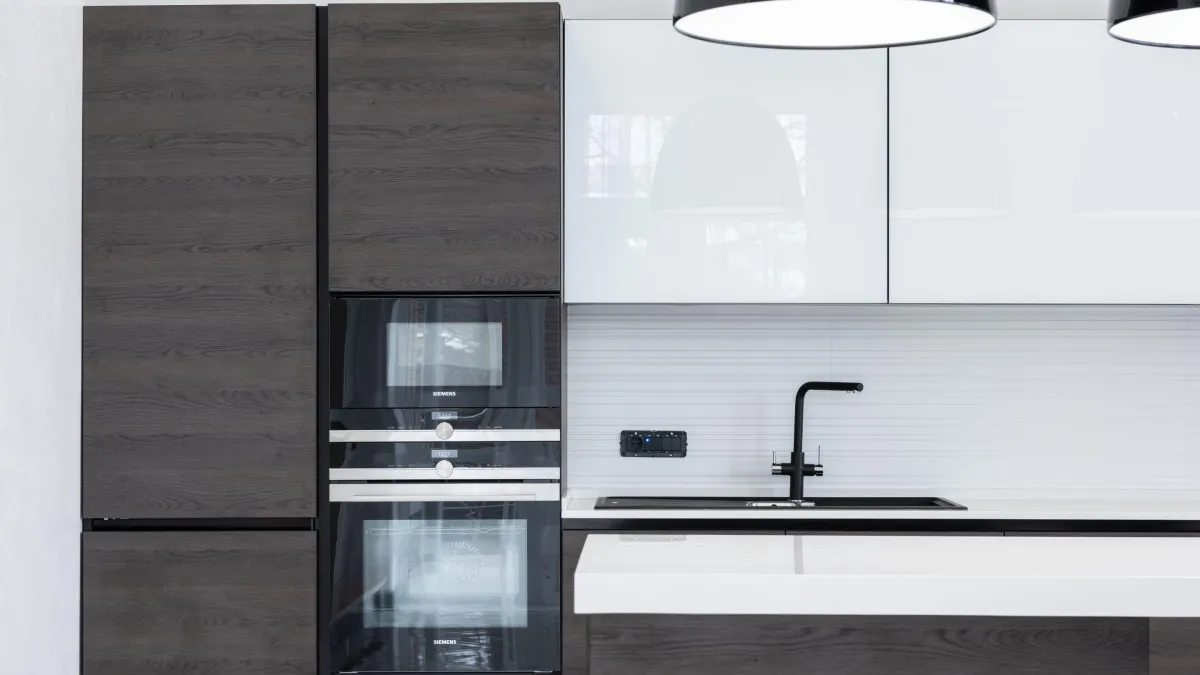 This image captures the essence of a modern kitchen with its clean lines and minimalist design. Dark, wood-grain cabinetry offers a sophisticated contrast to the sleek white overhead units. The high-tech Siemens appliances, including an oven and a wine cooler, are built seamlessly into the cabinetry, exemplifying a smart use of space and cutting-edge design. The monochromatic color scheme is punctuated by the matte black faucet and cooktop, reinforcing the contemporary feel. Overhead, the simple yet elegant pendant lights add a soft illumination to the space. This kitchen is a testament to innovative design where every detail contributes to a functional yet aesthetically pleasing culinary environment.