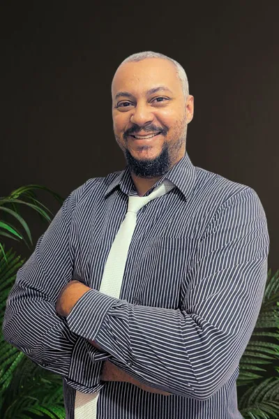 Valdemire Tavares Smiling with his arms crossed in a striped button-up shirt and white tie. Standing in front of a brow background with green plants behind him.