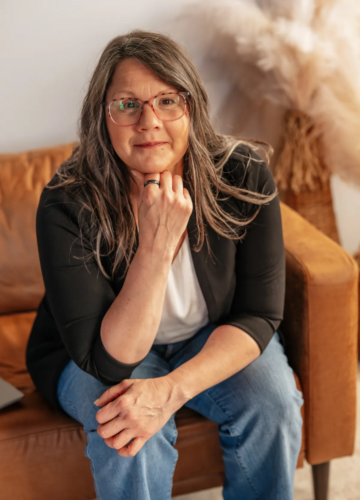Brunette woman - Stacey Feidt smiling in a white shirt, black blazer, and blue jeans. Sitting on a tan couch.