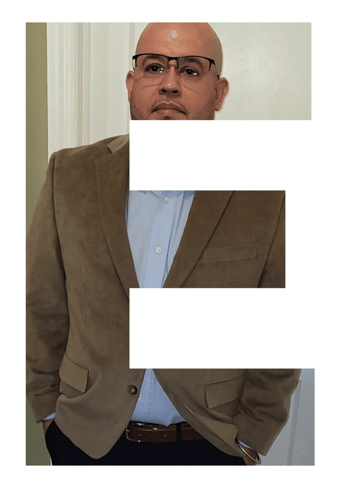 Eufemio Perez wearing glasses and a brown blazer over a light blue button-down shirt, standing indoors with hands in his pockets and a neutral expression.