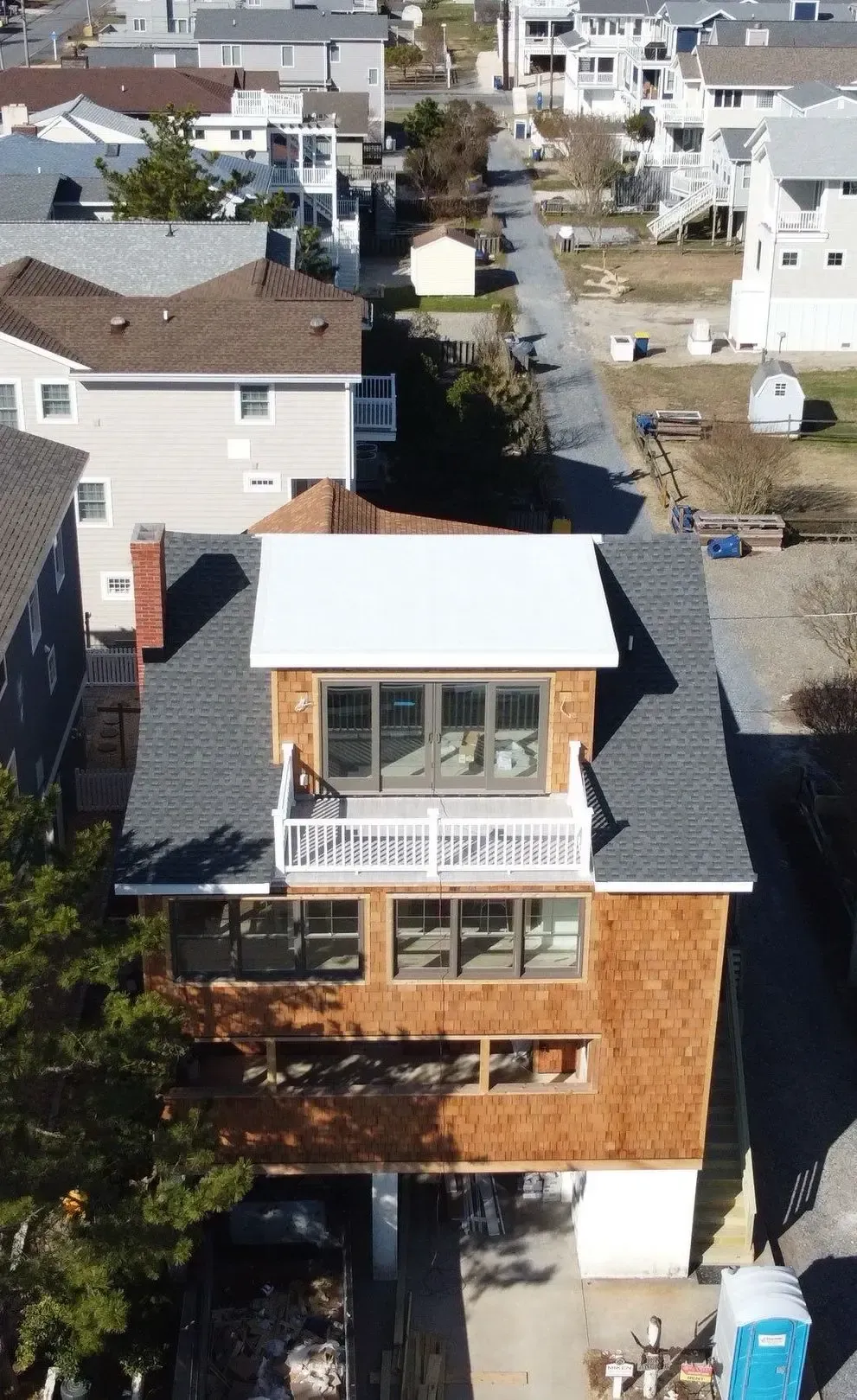 Aerial view of a coastal residence with dark asphalt shingles, a white low-slope roof, balconies, and neighboring homes.