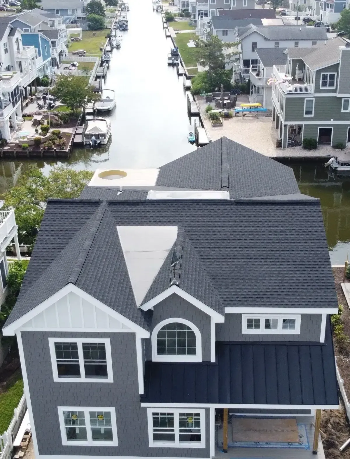 Aerial view of a blue-gray coastal home with a steep asphalt-shingle roof and a canal behind the property.