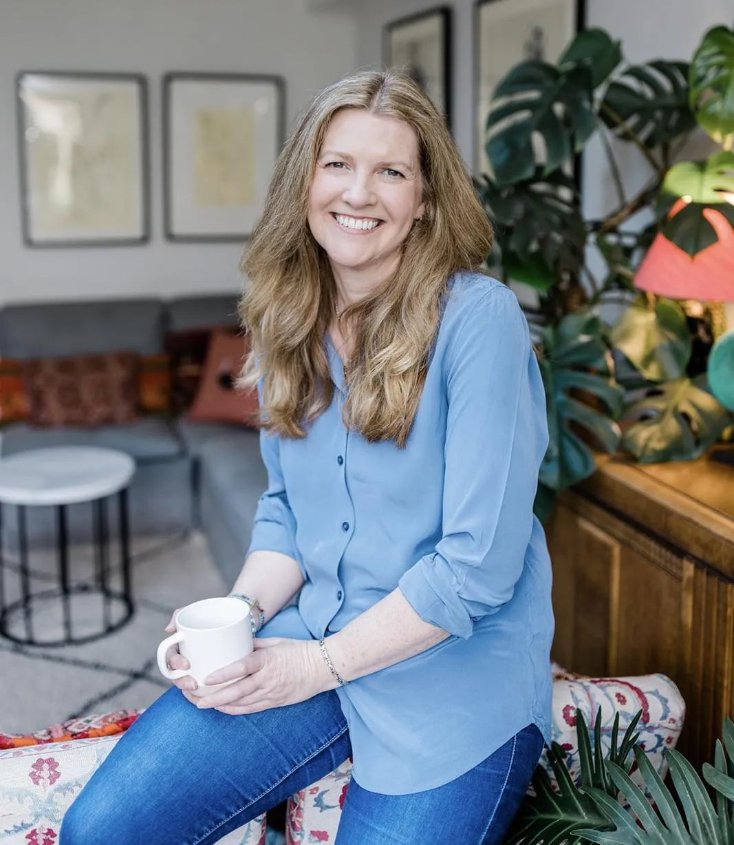 Suzi wearing blue jeans and a blue shirt perched on the back of a couch , smiling and holding a cup of tea.