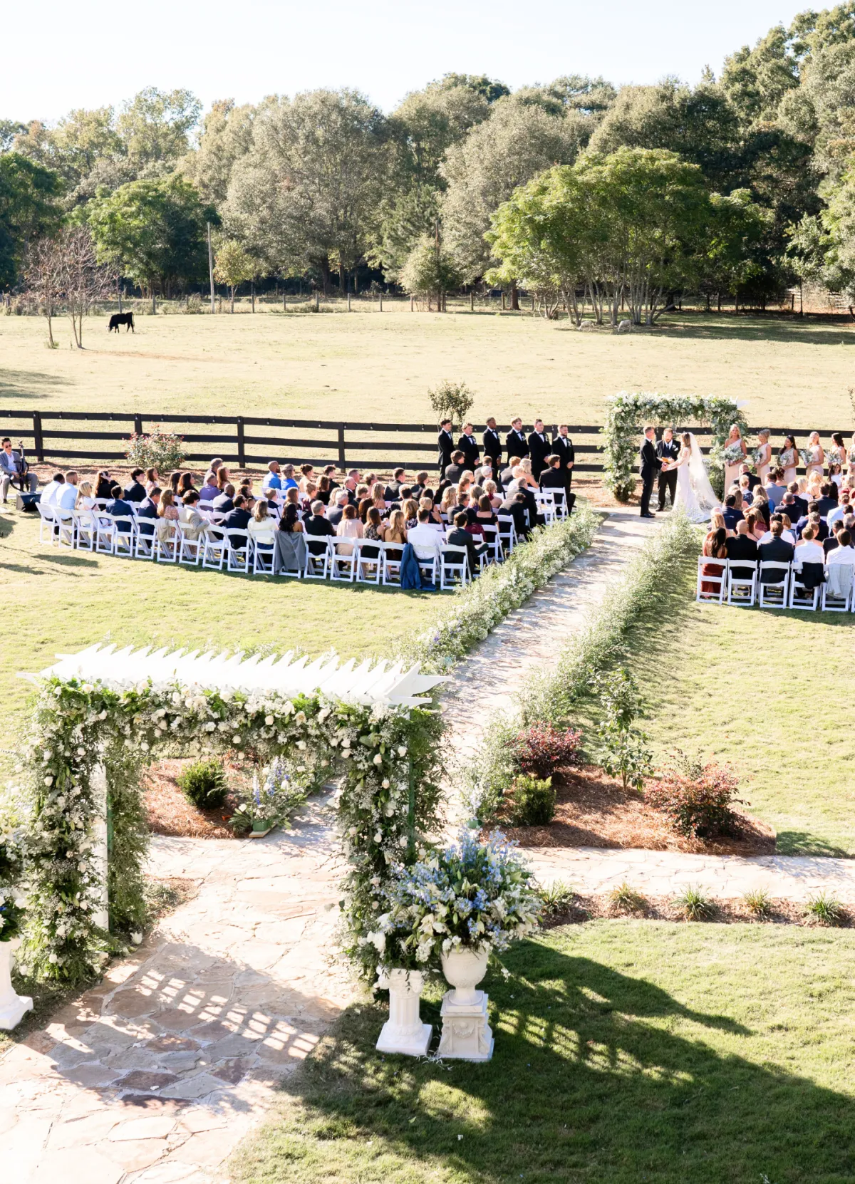 Outdoor wedding ceremony on the lawn at The Grove Estate, guests seated along a stone aisle facing a white floral arch