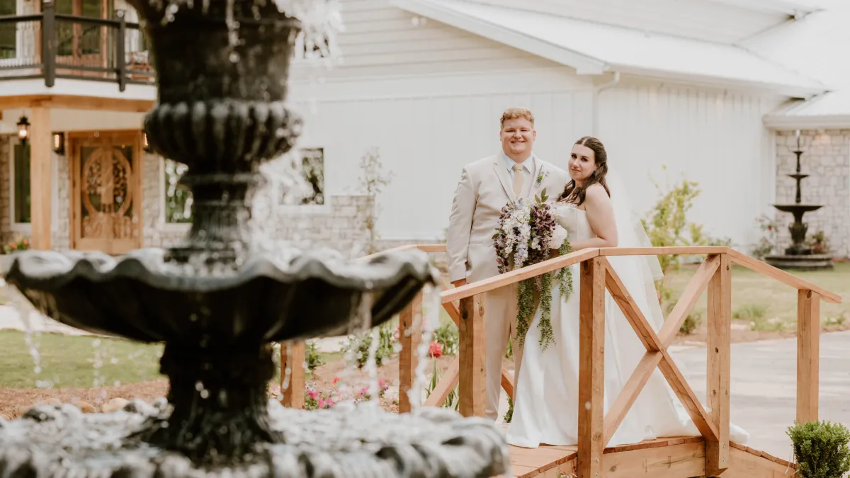 Bride and groom on the wooden footbridge outside The Grove Estate, with the stone building and tiered fountain behind them