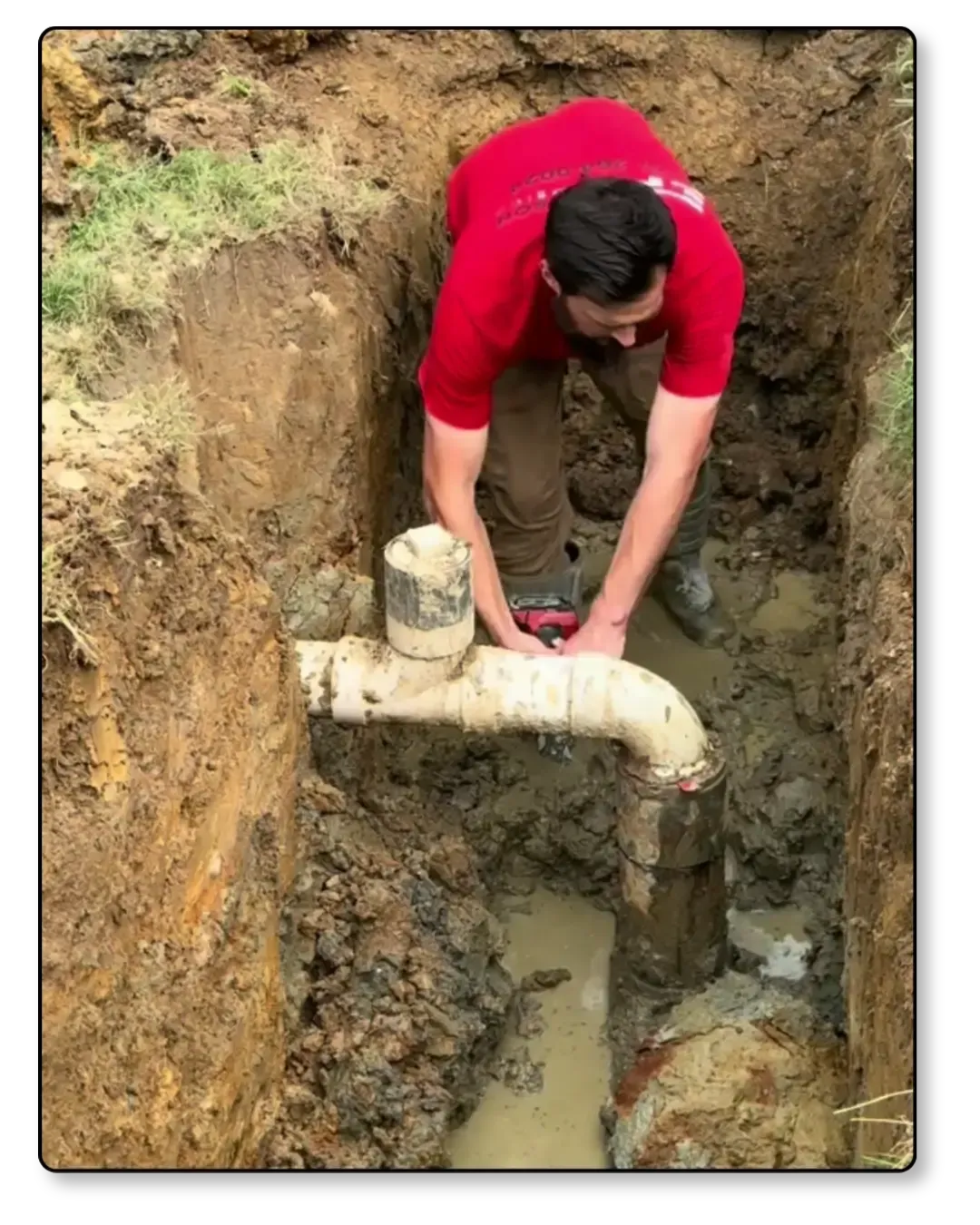 Person working in a muddy trench beside exposed white pipes.
