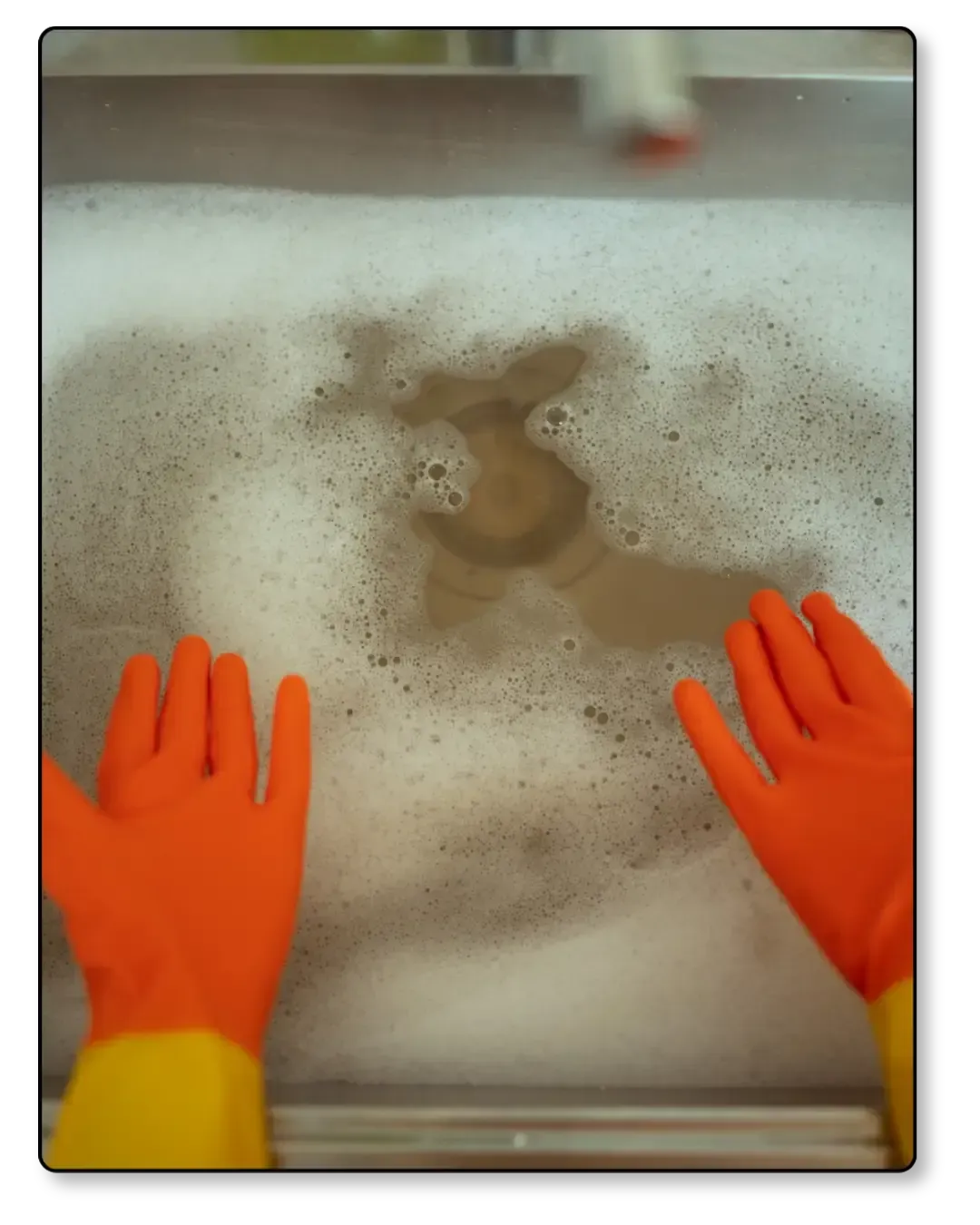 Hands in orange rubber gloves over a sink filled with soapy water.