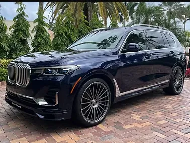 A white yacht docked at a marina, with a detailer polishing the hull under clear blue skies, and an RV parked nearby, both gleaming and surrounded by water and palm trees.