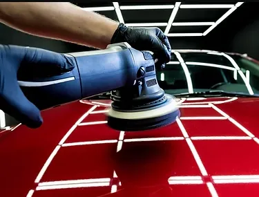 A technician using a dual-action polisher on a deep blue sedan, with dramatic before-and-after reflections visible on the hood, set in a well-lit, professional detailing bay.