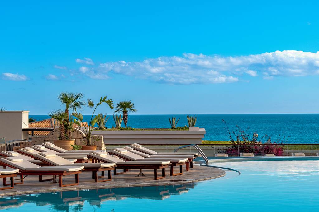 Luxury sunbeds sit next to a pool against a backdrop blue sea and sky in Greece