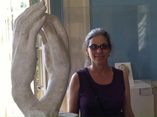 Frederick, MD CranioSacral Therapist Lori Leitzel Rice, haired pulled back and in a black tank top, stands next to a sculpture of two hands coming together