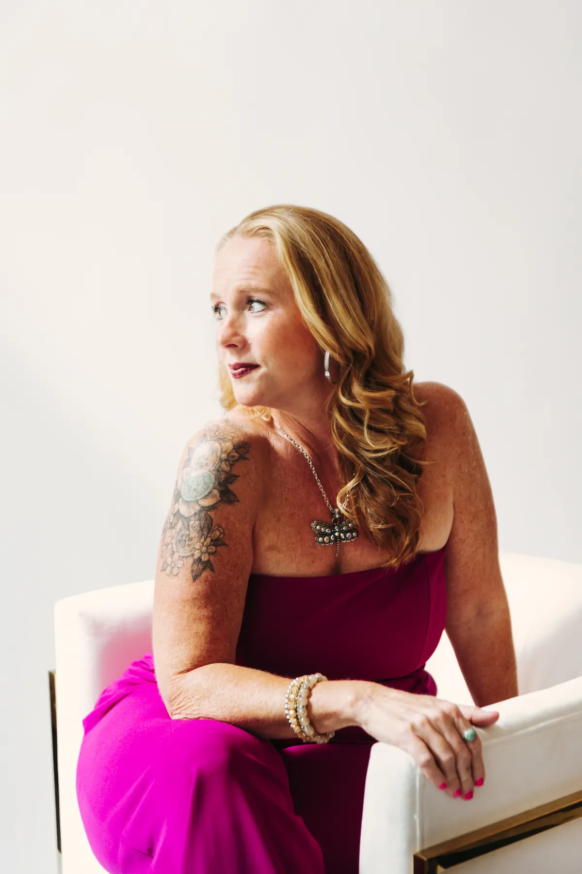 Becca Powers® seated in a white chair in a fuchsia jumpsuit, gazing to the side