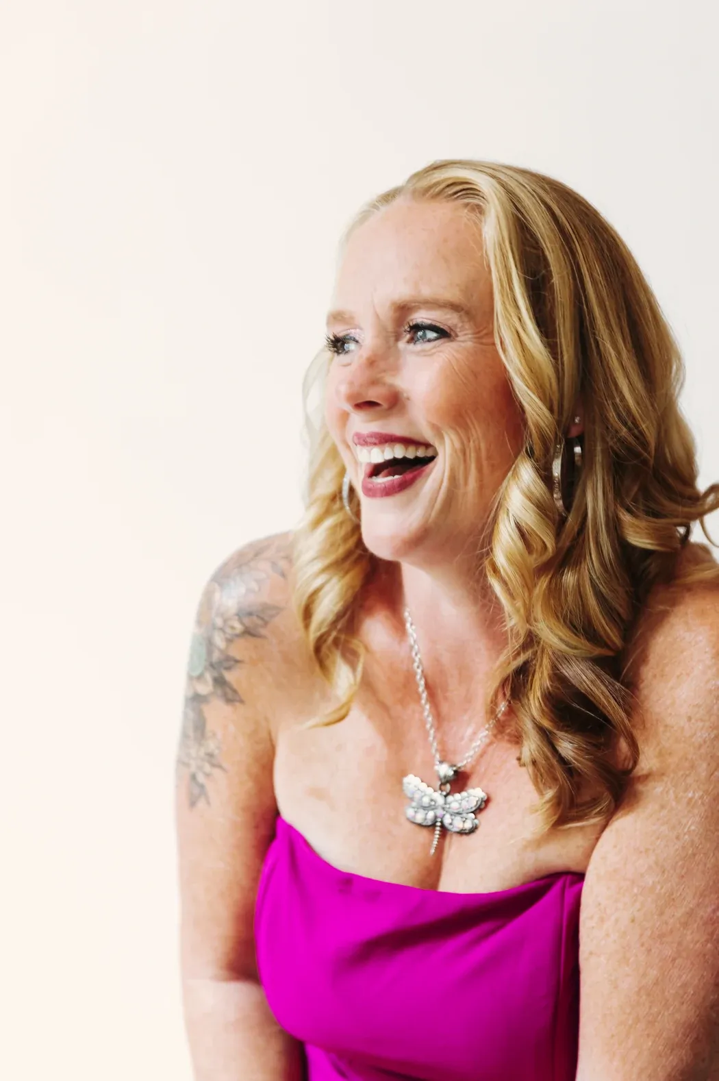 Becca Powers® laughing in a strapless fuchsia top against a white backdrop, turned to the side