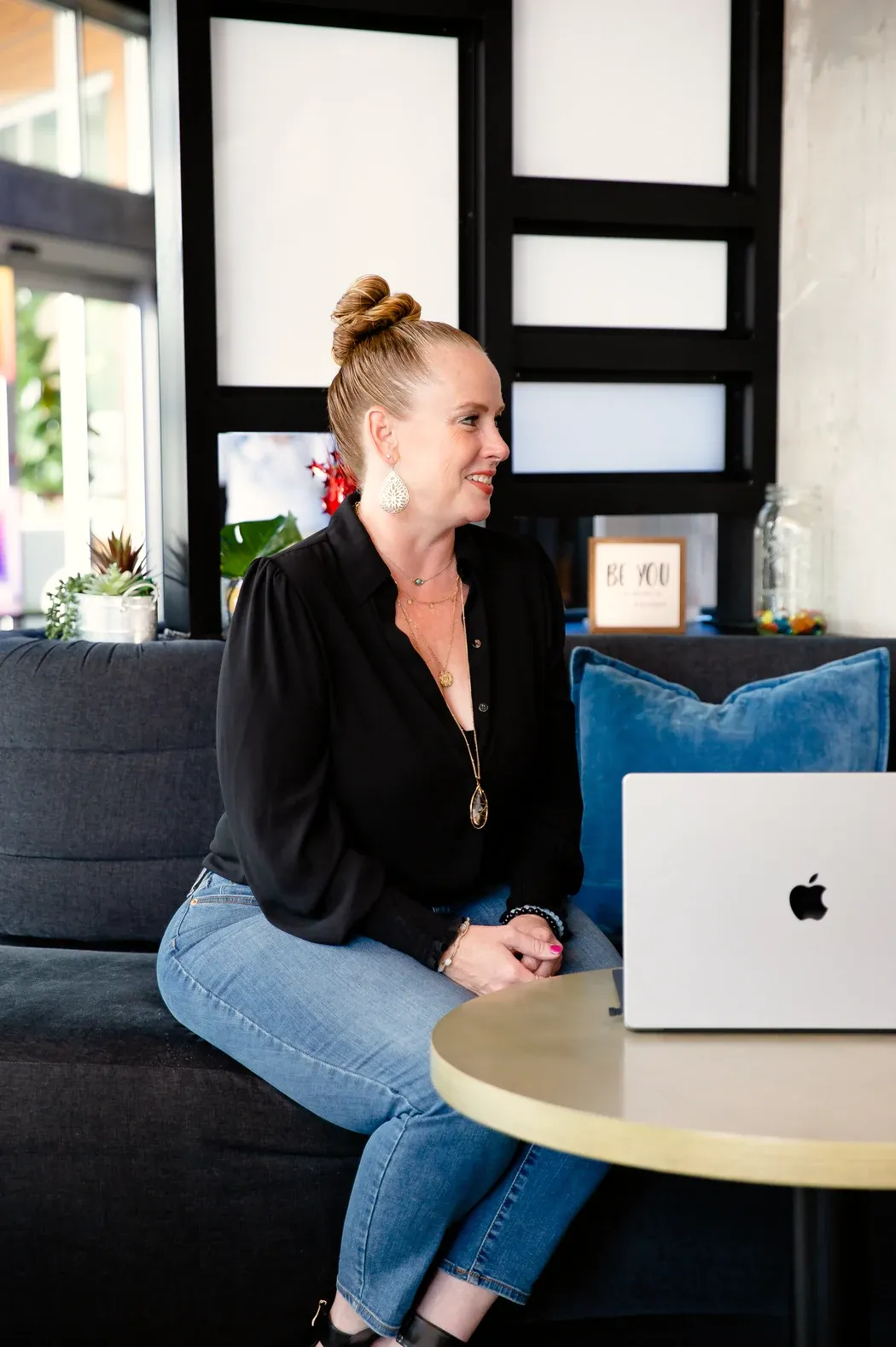 Becca Powers® seated on a lounge sofa beside her laptop, smiling as she looks toward the conversation