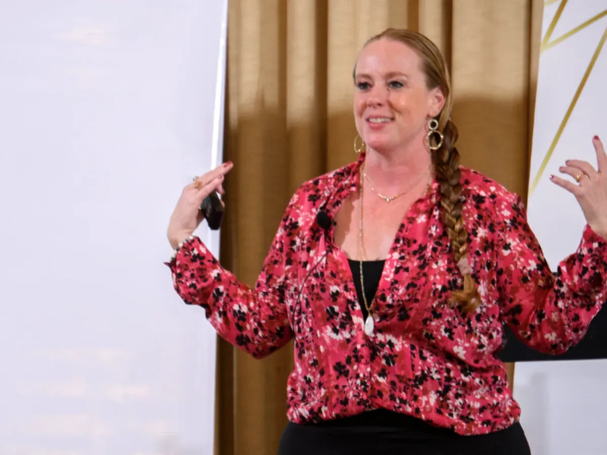 Becca Powers® speaking on stage in a red floral blouse, for booking her as a keynote speaker