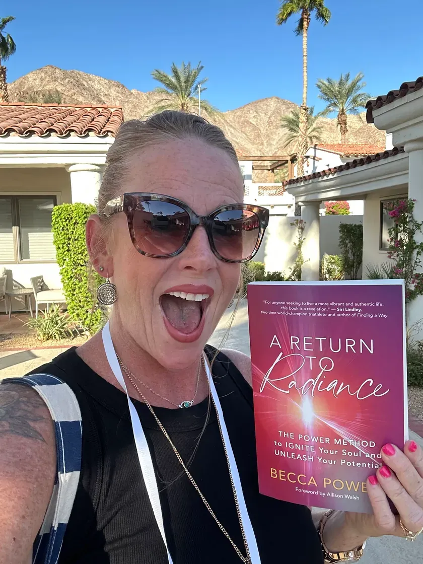 Becca Powers® outdoors among palm trees in sunglasses, cheering as she holds up her book A Return to Radiance