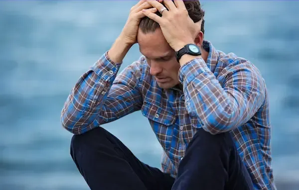 Man sitting by the water with his head in his hands, overwhelmed and reflecting during a relationship crisis—representing men’s relationship coaching with Vanessa Cardenas.
