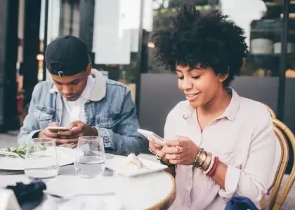 Photo of a couple sitting together in a public space while distracted by devices, representing emotional disconnection and loss of shared relational space