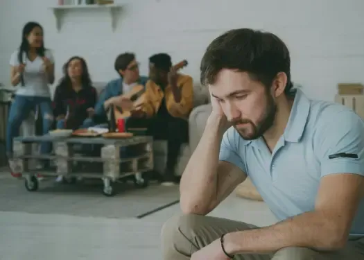 Man sitting alone in a room while others socialize in the background, representing emotional isolation in relationships