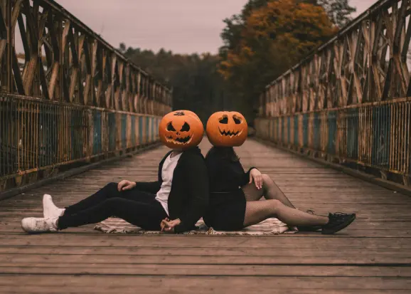 Couple wearing jack-o'-lantern pumpkin masks sitting back-to-back on a wooden bridge while holding hands, symbolizing connection and emotional distance in relationships.