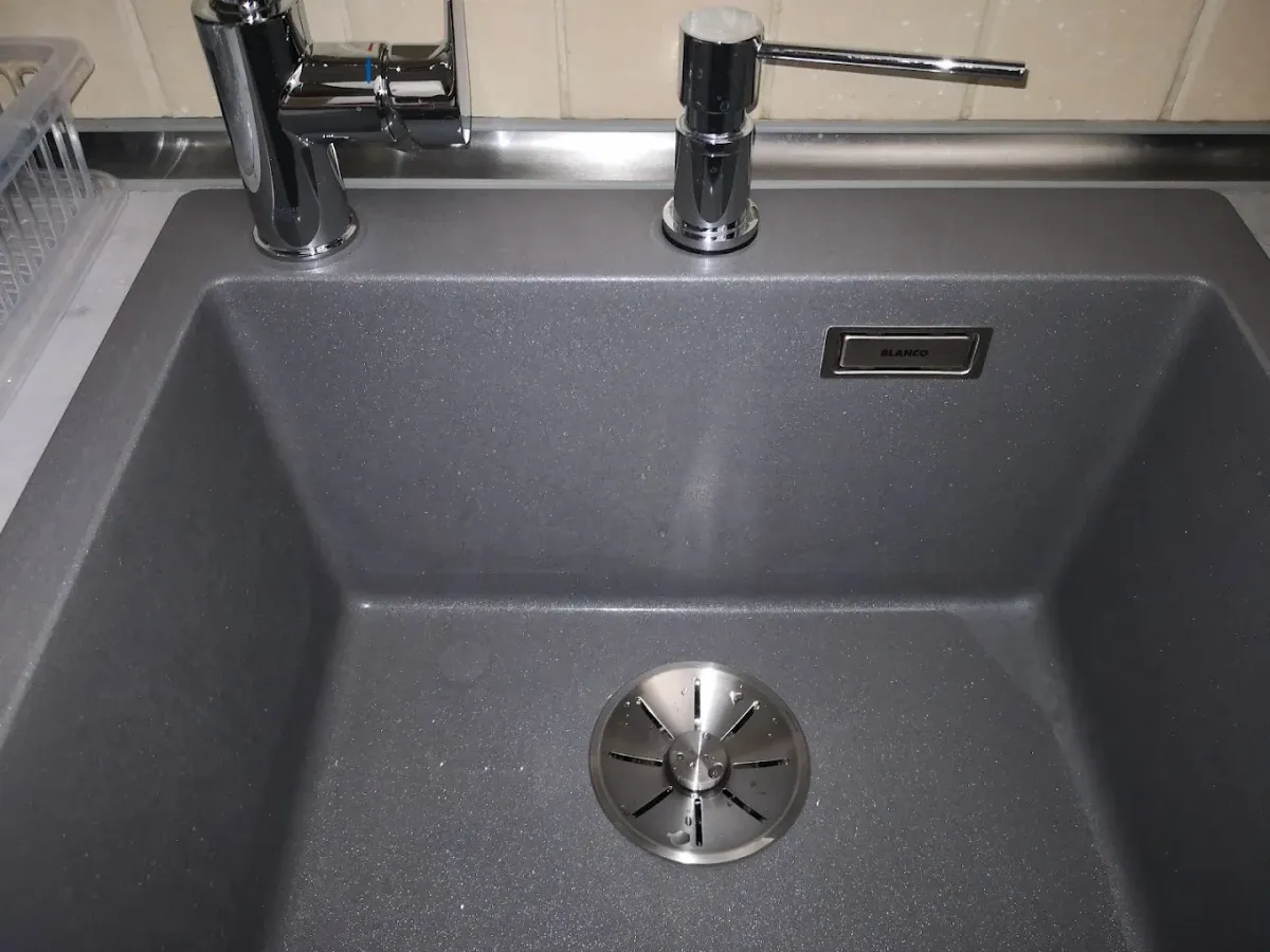 Sötétszürke, gránithatású konyhai mosogatótálca felülnézeti képe. A tálca bal oldalán egy elegáns, krómozott Grohe csaptelep, mellette jobbra pedig egy beépített krómozott szappanadagoló található. A medence alján egy szálcsiszolt fém lefolyószűrő, a hátfalon pedig egy szögletes "BLANCO" feliratú túlfolyó takaróelem látszik.