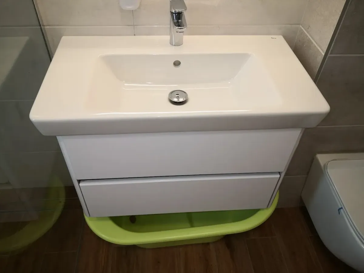 Egy modern fürdőszobai mosdószekrény látható. A falra szerelt, fehér, kétfiókos szekrény tetején egy szögletes, széles peremű fehér kerámia mosdókagyló (Roca) ül, rajta krómozott egykaros csapteleppel. A szekrény alatt, a famintás padlócsempén egy neonzöld műanyag babakád van betolva. A háttérben világos falicsempék láthatók.