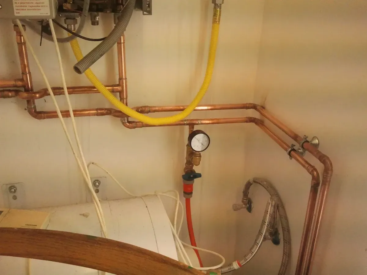 Egy kazán vagy vízmelegítő alatti csőhálózat látható a sarokban. Vörösréz csövek futnak vízszintesen és függőlegesen, forrasztott idomokkal. Egy sárga bordázott flexibilis gázcső is húzódik a fal mentén. Lent egy nyomásmérő óra (manométer) látszik, amelyhez egy kék karos csappal ellátott piros cső csatlakozik. Alul egy fehér tartály (bojler) teteje tűnik fel.