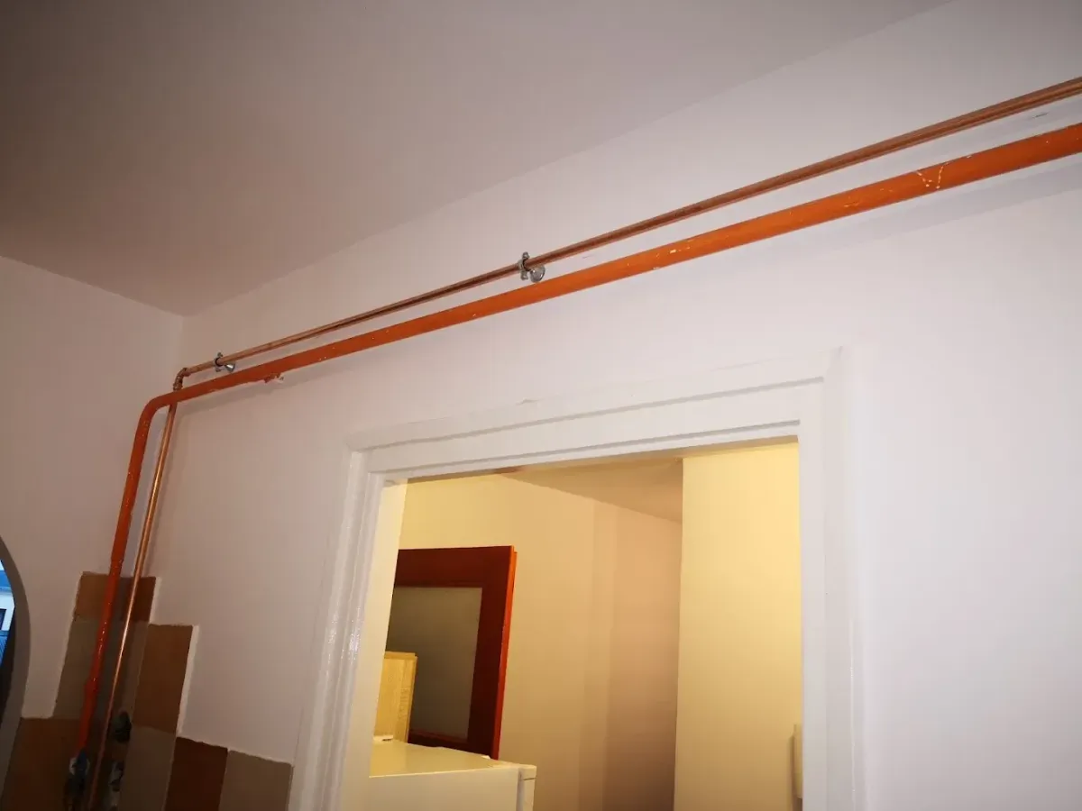 Egy szoba fehérre festett sarka, ahol egy vastag, narancssárgára festett fémcső (valószínűleg gázcső) fut vízszintesen a mennyezet alatt, majd 90 fokban lefelé kanyarodik a fal mentén. A csövet fém bilincsek rögzítik. Középen egy nyitott fehér ajtókeret látható, amely egy másik helyiségbe vezet. A bal alsó sarokban barna és bézs falicsempék részlete is feltűnik.