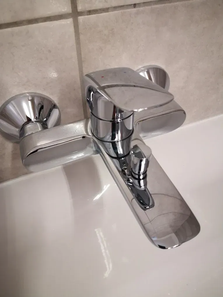 Robusztus, modern dizájnú fali kádcsaptelep csillogó króm kivitelben. A széles, lapos keverőkar és az elnyújtott kifolyó dominálja a képet, utóbbin egy hengeres zuhanyváltó gomb is helyet kapott. A csap két kerek fémrózsával simul a bézs árnyalatú csempézett falhoz, alatta pedig egy hófehér kád vagy szaniter pereme veri vissza a fényt.