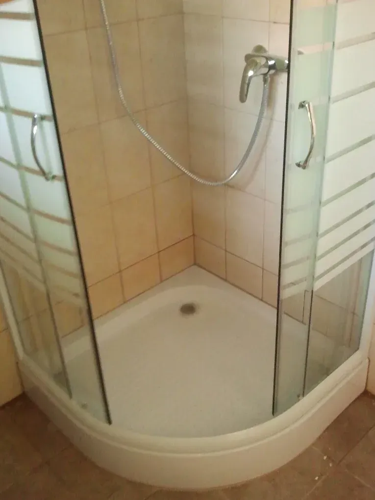 Egy fürdőszobai sarok zuhanykabin látható fehér, íves zuhanytálcával. A kabin üvegajtói vízszintesen csíkozottak (homokfúvott hatásúak) és krómozott fém fogantyúkkal vannak felszerelve. A belső falakat világosbarna, négyzet alakú csempék borítják, a falon egy krómozott zuhanycsaptelep és gégecső lóg.