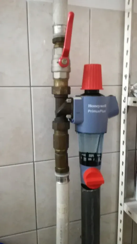 Függőleges vízvezeték-szerelvények láthatók fehér csempés fal előtt. Középen sárgaréz idomok és egy piros karos golyóscsap található. Jobb oldalon egy kék színű, Honeywell PrimusPlus márkájú vízszűrő és nyomáscsökkentő állomás van felszerelve, felül és alul piros műanyag tekerőgombokkal, átlátszó szűrőházzal.