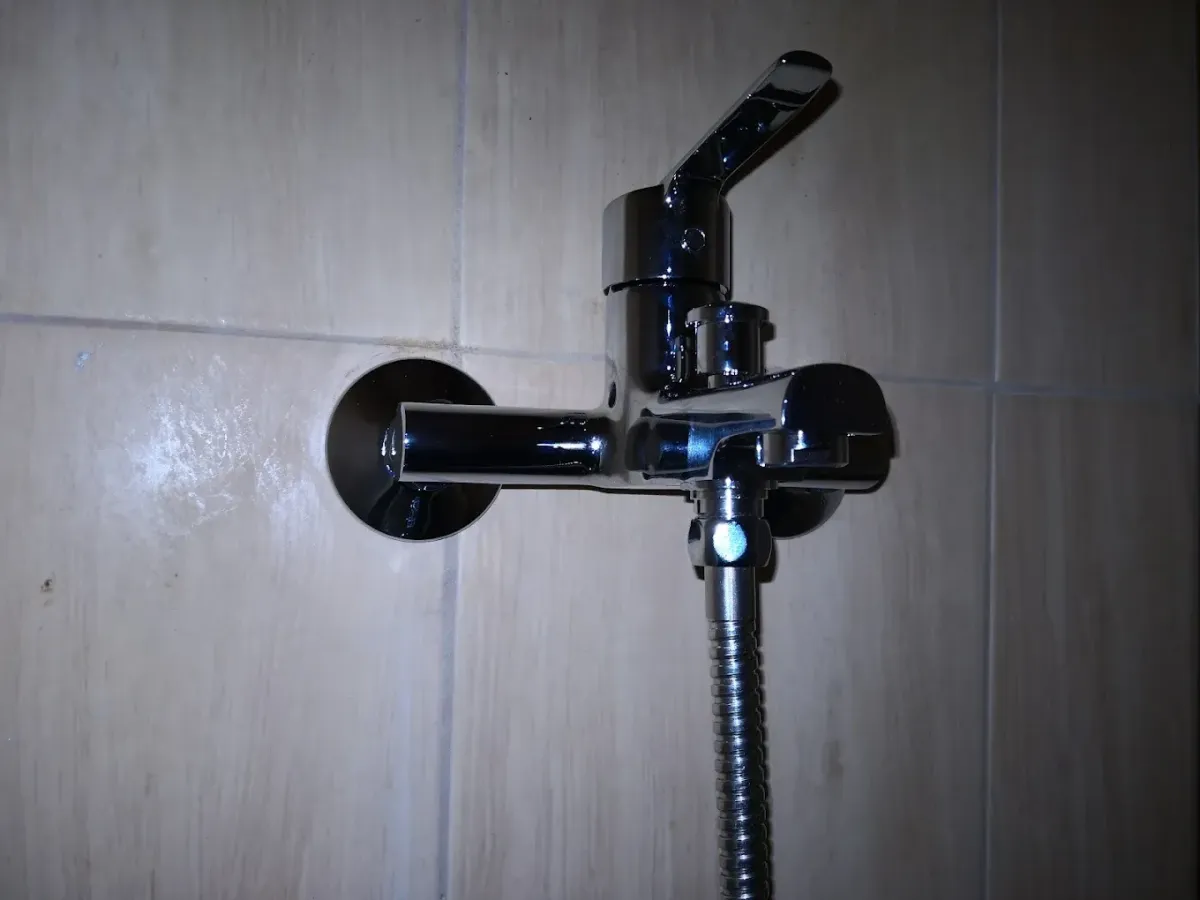 Közeli felvétel egy krómozott fali kád- és zuhanycsaptelepről, világos bézs színű falicsempére szerelve. A csap egykaros, rövid kifolyóval és egy felhúzható zuhanyváltó gombbal rendelkezik. Alulról egy fém spirálos gégecső csatlakozik hozzá.