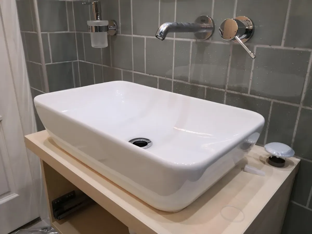 Egy elegáns fürdőszoba részlete, fókuszban egy pultra ültetett, téglalap alakú, lekerekített sarkú fehér kerámia mosdókagylóval. A mosdó egy világos, fahatású pulton áll, amelynek fiókja félig nyitva van, felfedve a sínrendszert. A falra egy modern, krómozott Grohe csaptelep van beépítve, különálló kifolyóval és keverőkarral. A hátteret szürkészöld, négyzet alakú csempék adják, balra pedig egy fali szappanadagoló látszik.