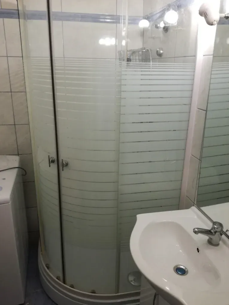 Egy fürdőszoba részlete látható, amelynek fókuszában egy íves sarok zuhanykabin áll, vízszintes csíkozású, félig átlátszó üvegajtókkal. A zuhanykabin mellett jobbra egy fehér kerámia mosdókagyló található krómozott csapteleppel, a bal oldalon pedig egy fehér mosógép széle lóg a képbe. A falakat világos csempe borítja, felül egy kék díszítőcsíkkal.