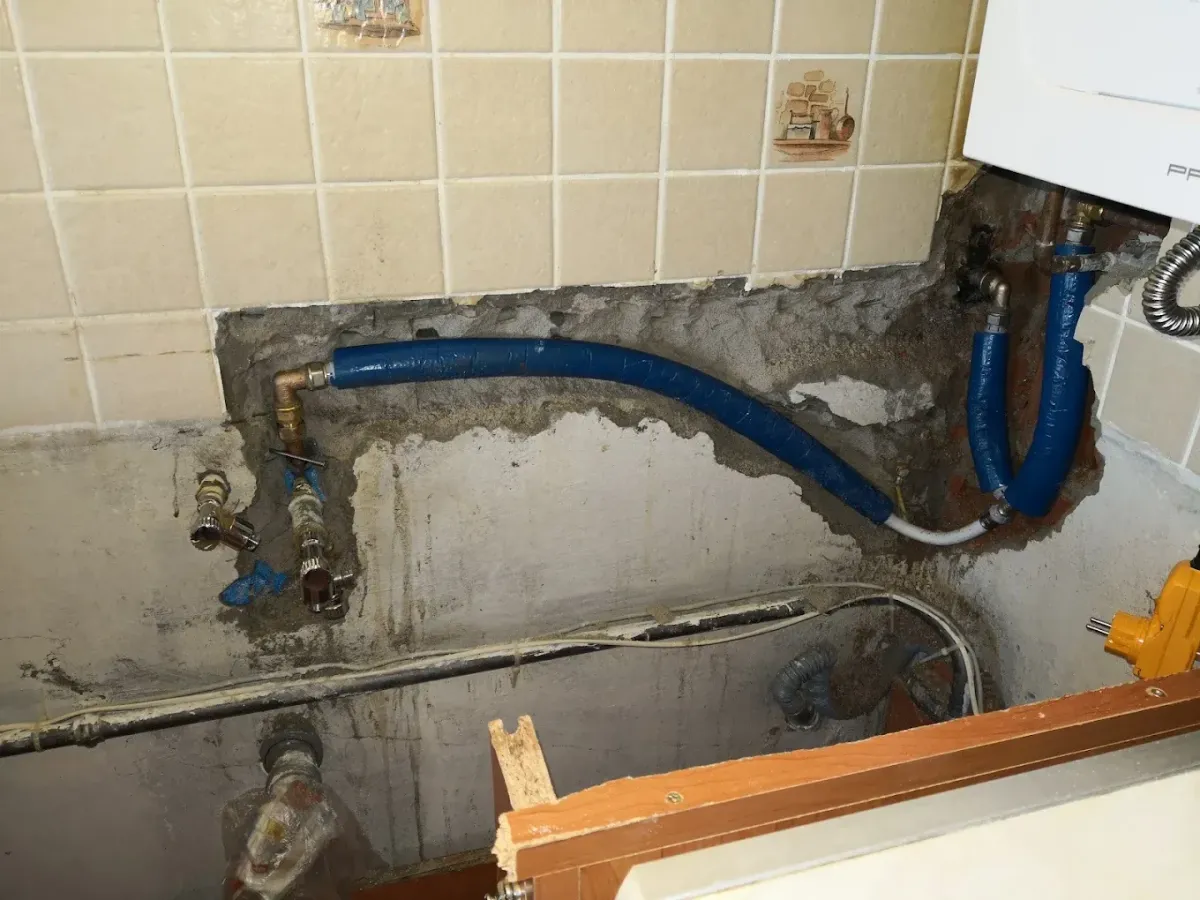 Egy felújítás alatt álló fürdőszoba vagy konyha fala látható, ahol a csempét és a vakolatot megbontották. A kibontott falrészben vastag, kék szigeteléssel bevont vízvezetékcsövek futnak, amelyek sárgaréz idomokkal csatlakoznak egymáshoz. Lefelé, a csupasz betonfal mentén további régi csövek, vezetékek és lefolyók láthatók, valamint némi törmelék. A kép jobb szélén egy sárga elektromos szerszám egy része is feltűnik.