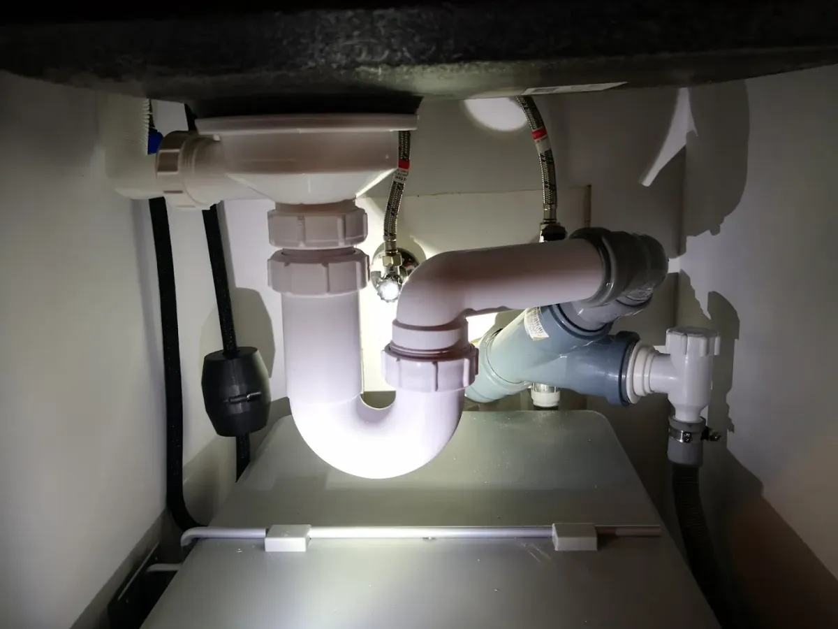 Konyhai mosogató alatti szerelvények láthatók sötét környezetben, zseblámpával vagy vakuval megvilágítva. Középen a mosogató fehér műanyag szifonja (bűzzárója) és lefolyócsöve ereszkedik le, amely egy Y-alakú, szürke PVC csatornacsőbe csatlakozik. Hátul a falból kiálló sarokszelepek és flexibilis fémcsövek (hideg-meleg víz) láthatók, valamint egy fekete ellensúly is lóg egy fekete csövön. A szerelvények egy szürke kuka vagy tárolódoboz felett helyezkednek el.