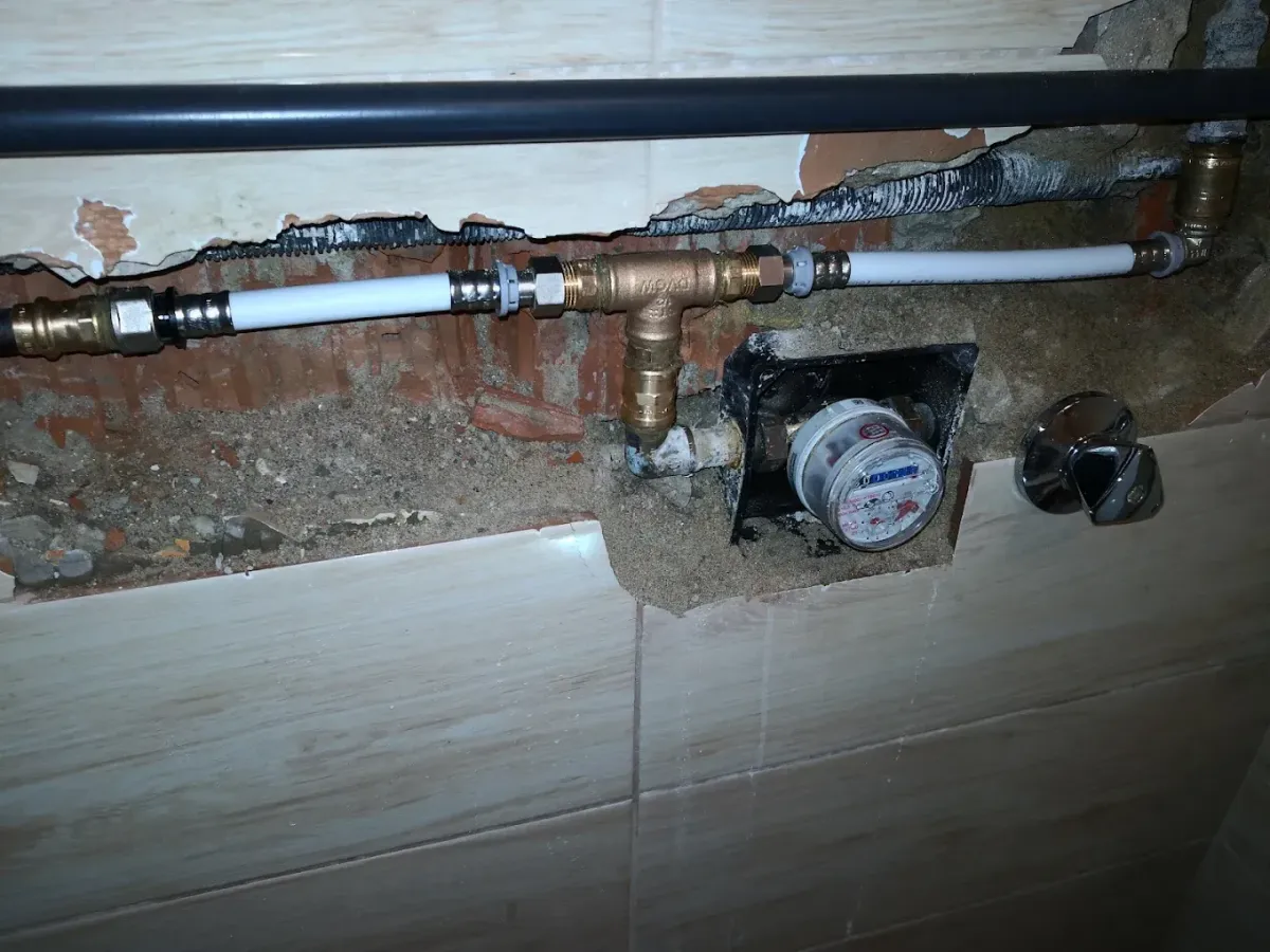 Egy felújítás alatt álló falrészlet, ahol a megbontott csempe és fal mögött vízszintesen futó fehér ötrétegű cső látható, réz csatlakozókkal és egy T-idommal. Ebből egy cső ágazik le egy falba süllyesztett analóg vízórához. A vízóra mellett jobbra egy krómozott elzárócsap van a még ép, famintás csempén.