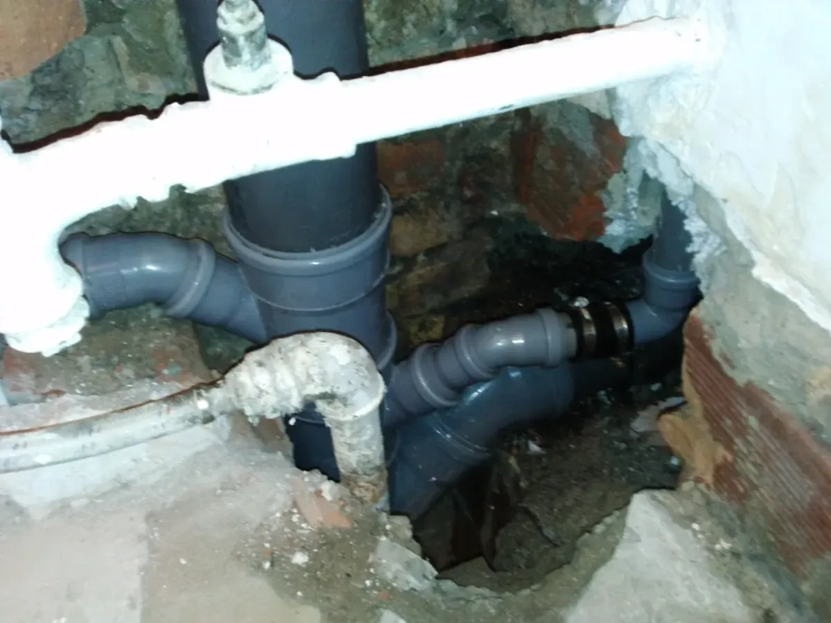 Gépészeti aknában vagy bontott falrészben lévő csőhálózat sötét felvétele. A háttérben vastag, szürke PVC lefolyócsövek futnak össze egy Y-idom segítségével, amelyhez egy fekete gumiszalagos bilincs is csatlakozik. Az előtérben felül egy vastag, fehérre festett cső keresztezi a képet, baloldalt pedig egy régebbi, koszos, kanyargó cső is látható a nyers tégla és törmelék között.